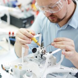 Man in protective glasses attaching action camera to quadcopter drone on table  with different tools in modern workshop